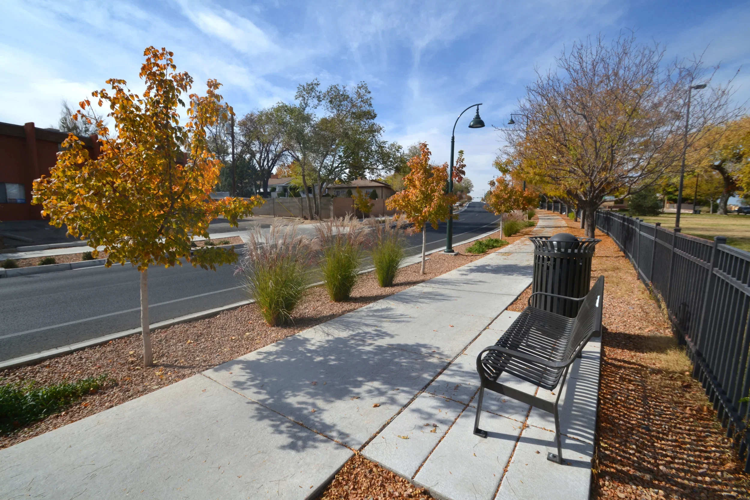 Empty sidewalk with trees showing fall foliage, a black bench, a trash can, and lampposts along a fenced park area, on a clear day.