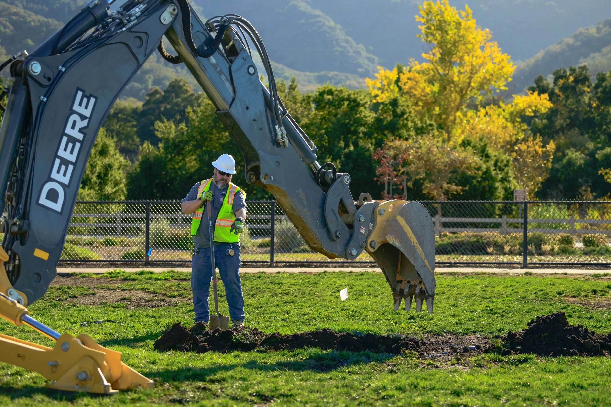 A construction worker in safety gear standing near a large excavator with the arm extended downward on a grassy area with trees and mountains in the background.