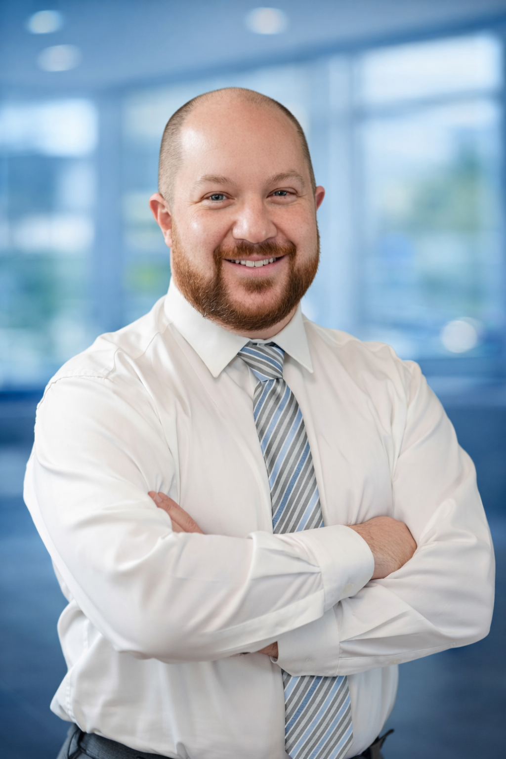 Smiling man with a beard in a white shirt and striped tie, arms crossed, standing in a modern office with large windows.