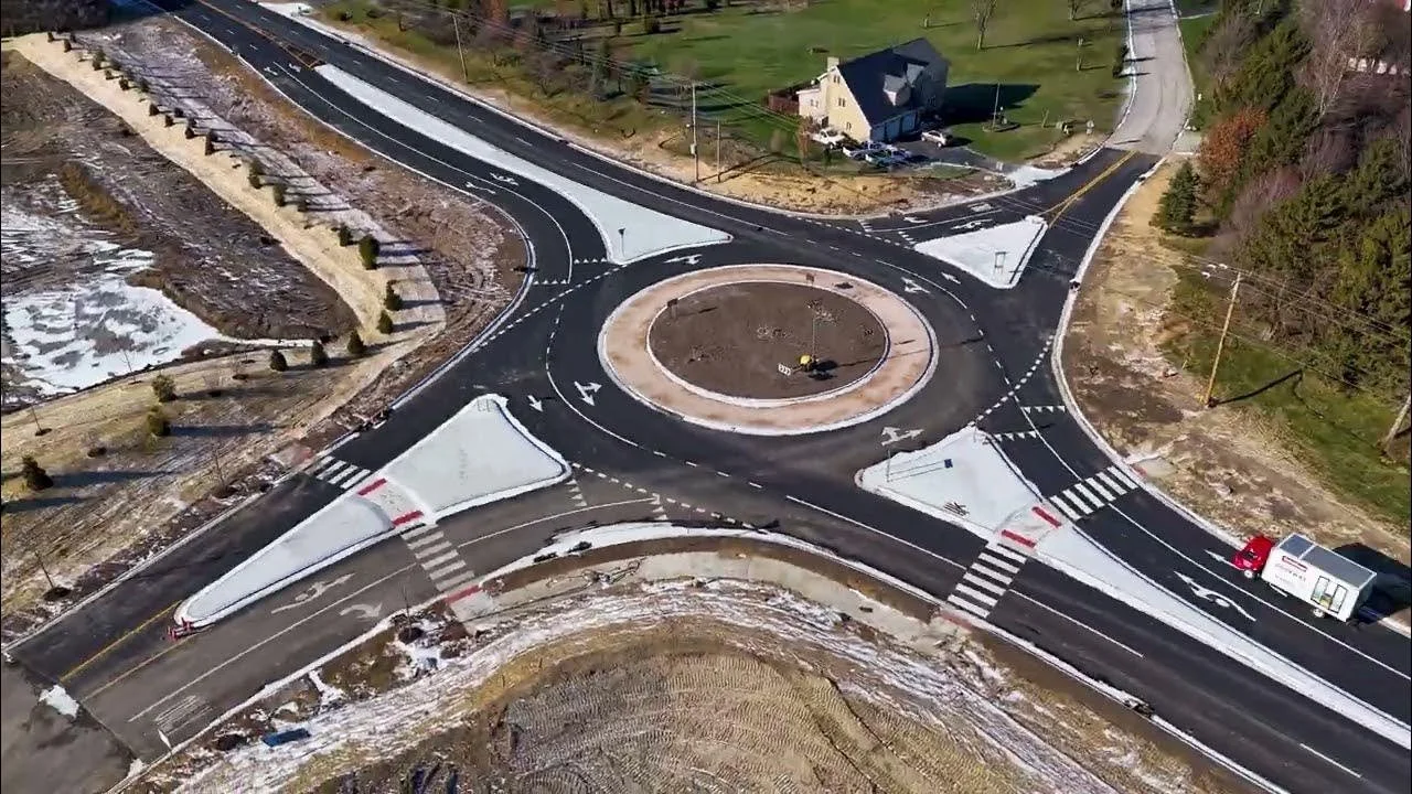 An aerial view of a new roundabout under construction at an intersection with crosswalks and traffic lanes, surrounded by trees and fields.