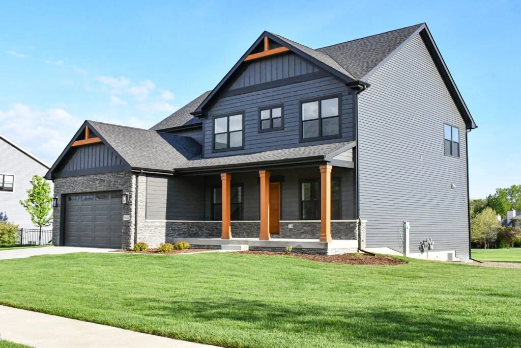 Modern two-story house with gray siding, stone accents, a garage, front porch with wooden columns, and lush green lawn.