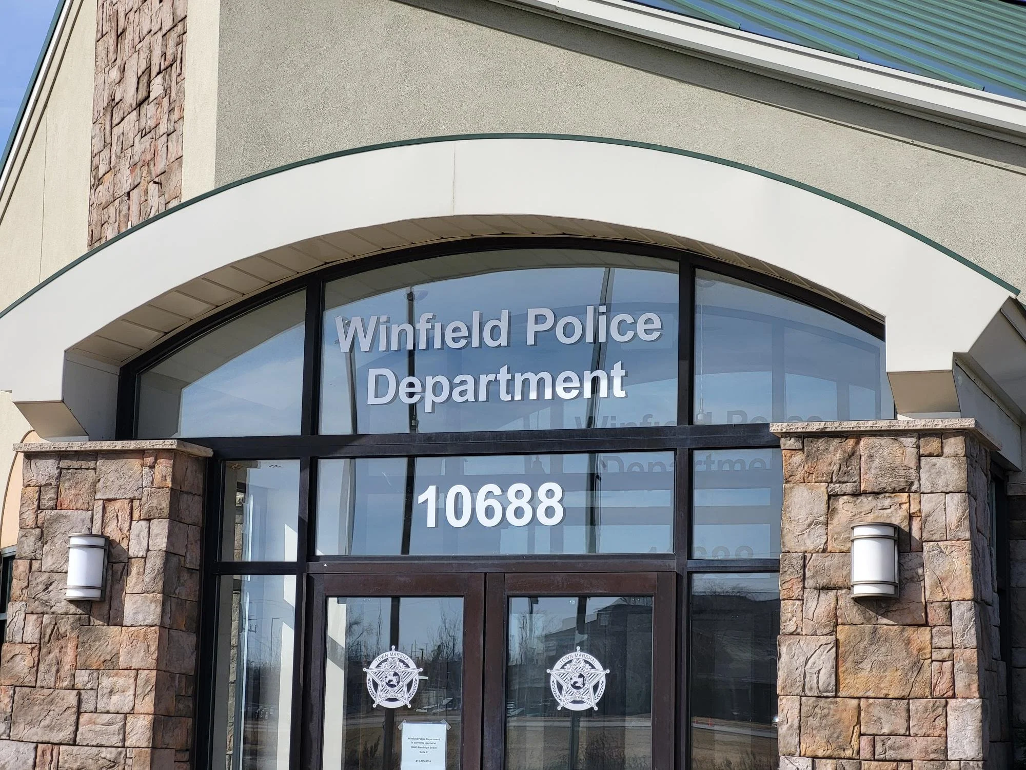 The front entrance of the Winfield Police Department building featuring large glass windows with the department's name and address, flanked by stone pillars with wall-mounted lights.