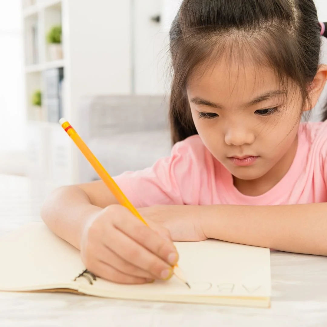 A young girl  learning to write at a young age in a notebook with a yellow pencil at a table in a bright montessori studio room.