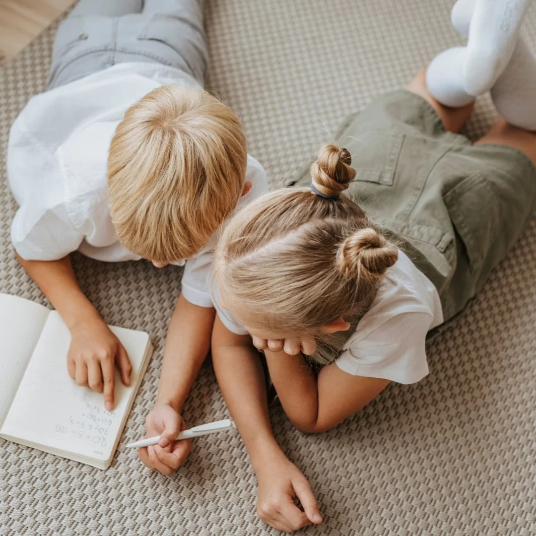 Two children lying on a checkered carpet, reading a book together and writing in a notebook. kids are enjoying learning at acton folsom.