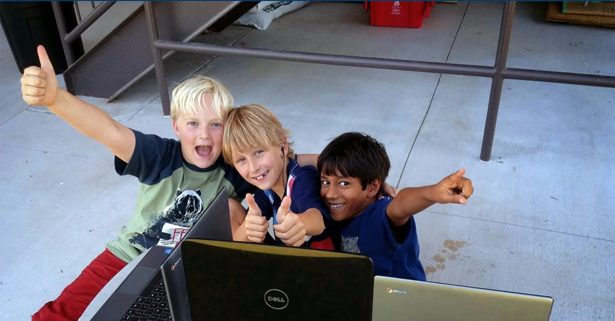 Three boys sitting on the ground outdoors, smiling and giving thumbs up, with laptops in front of them. they are working on core skills at their own pace. they love school in Folsom. 