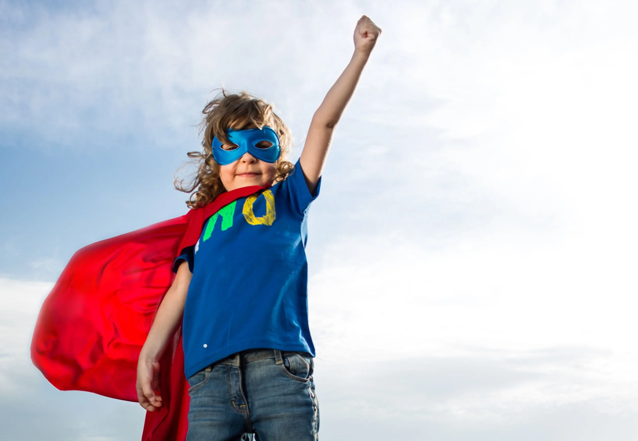 A young child dressed as a superhero, wearing a blue mask and a red cape, standing outdoors with a sky background, raising one fist in the air.