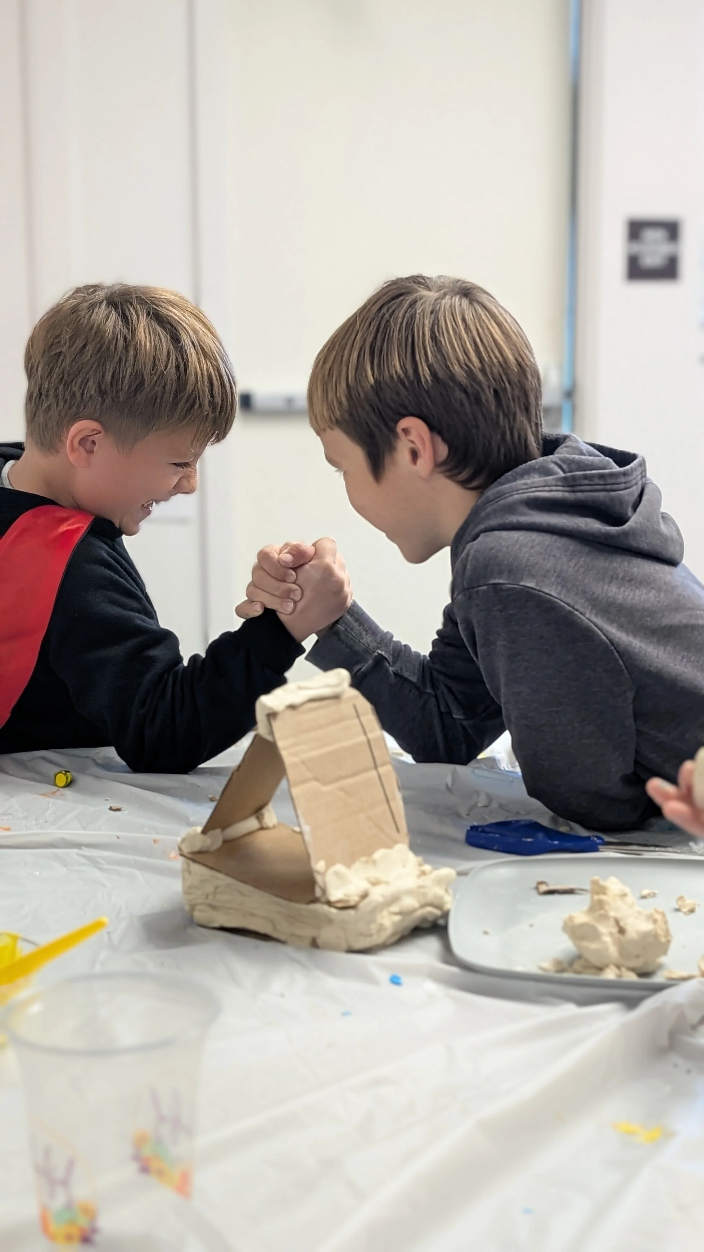 Two boys arm wrestling at a table with a white plastic tablecloth, surrounded by craft supplies, with a cardboard sculpture in front of them. the boys love school and challenges.