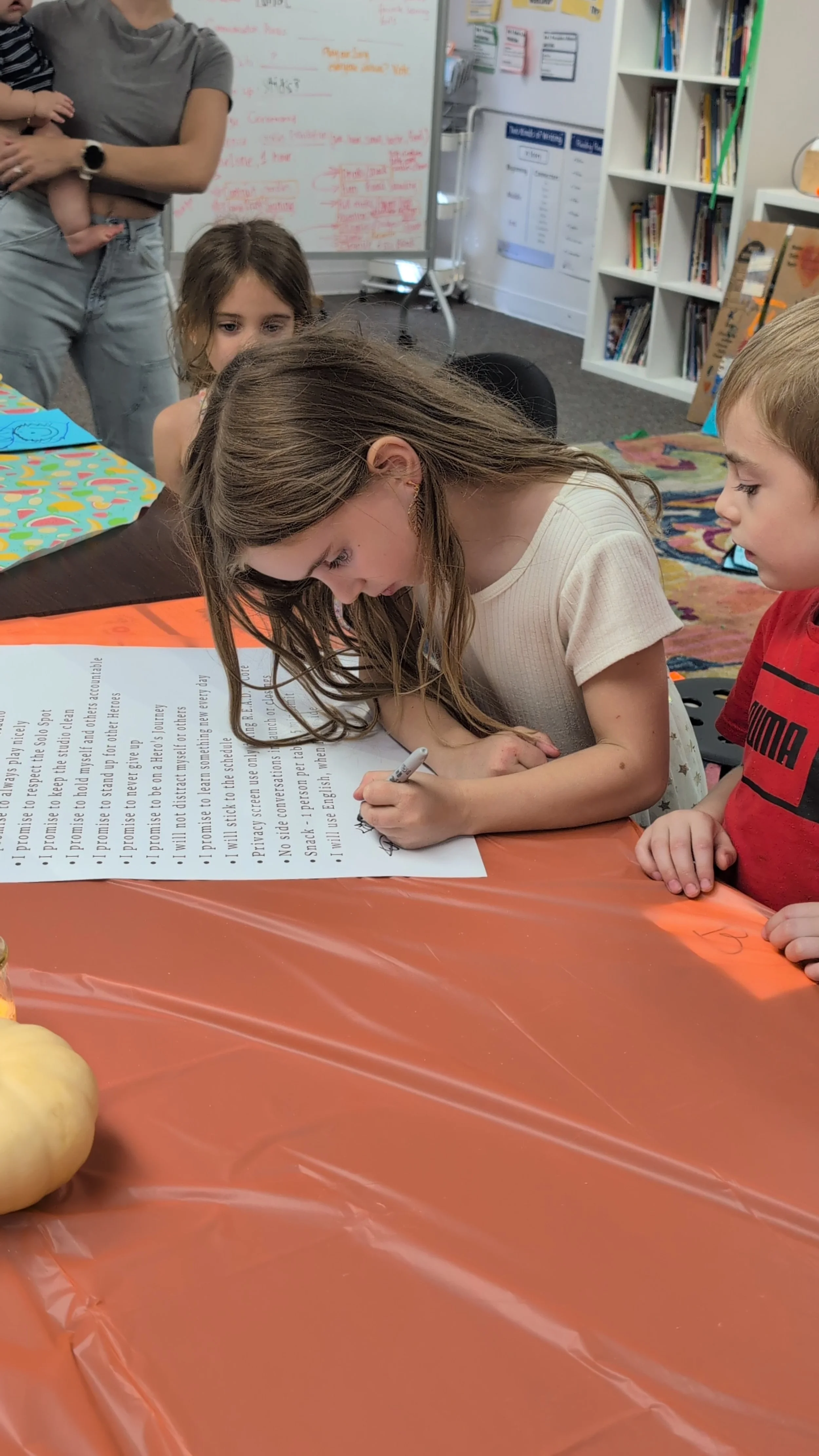 A young girl signing the studio contract that she helped put together. Other children watch, and a person holding a baby stands in the background. Bookshelves and a whiteboard with writing are visible in the classroom.