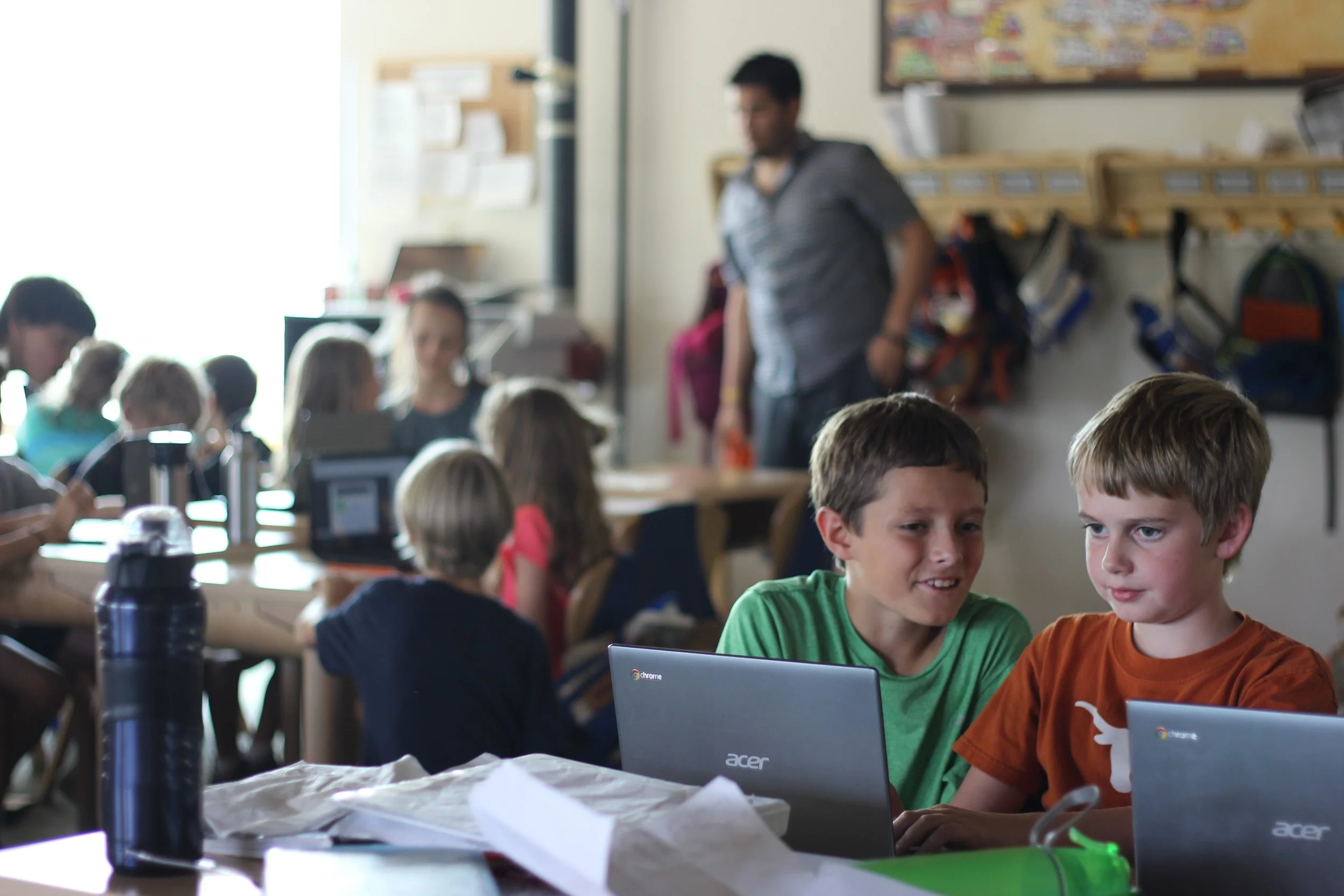 Two young boys sitting at a desk with laptops, engaged in a classroom activity, with other children and a teacher in the background.