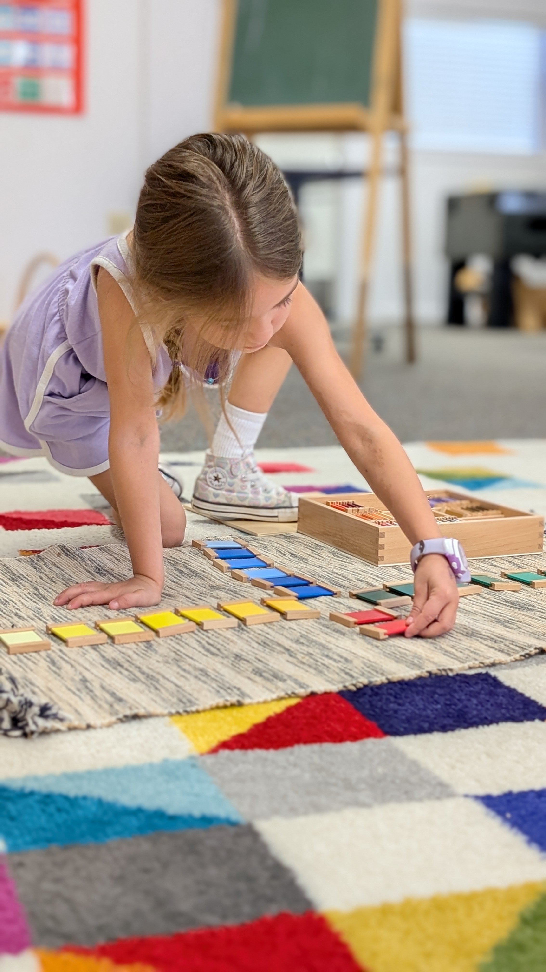 A young girl playing a game with colored tiles on a rug in a classroom, with a chalkboard and a window in the background. this is Sparks studio - an Acton Montessori studio with learner-led activities. 