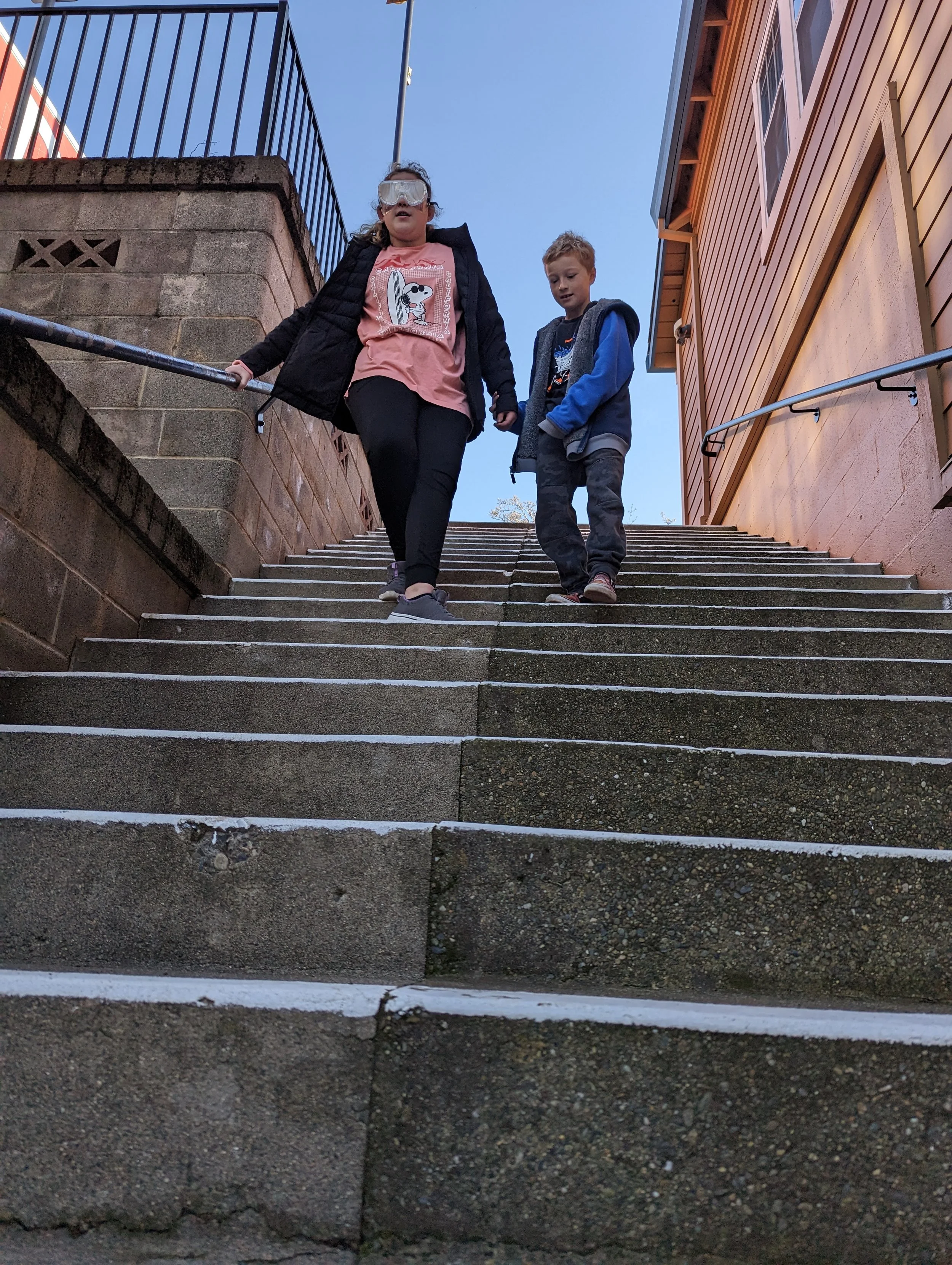 A girl and a young boy descend outdoor concrete stairs, with one child leading another in a project-led quest, where they experience real-word concepts at elementary age level. 