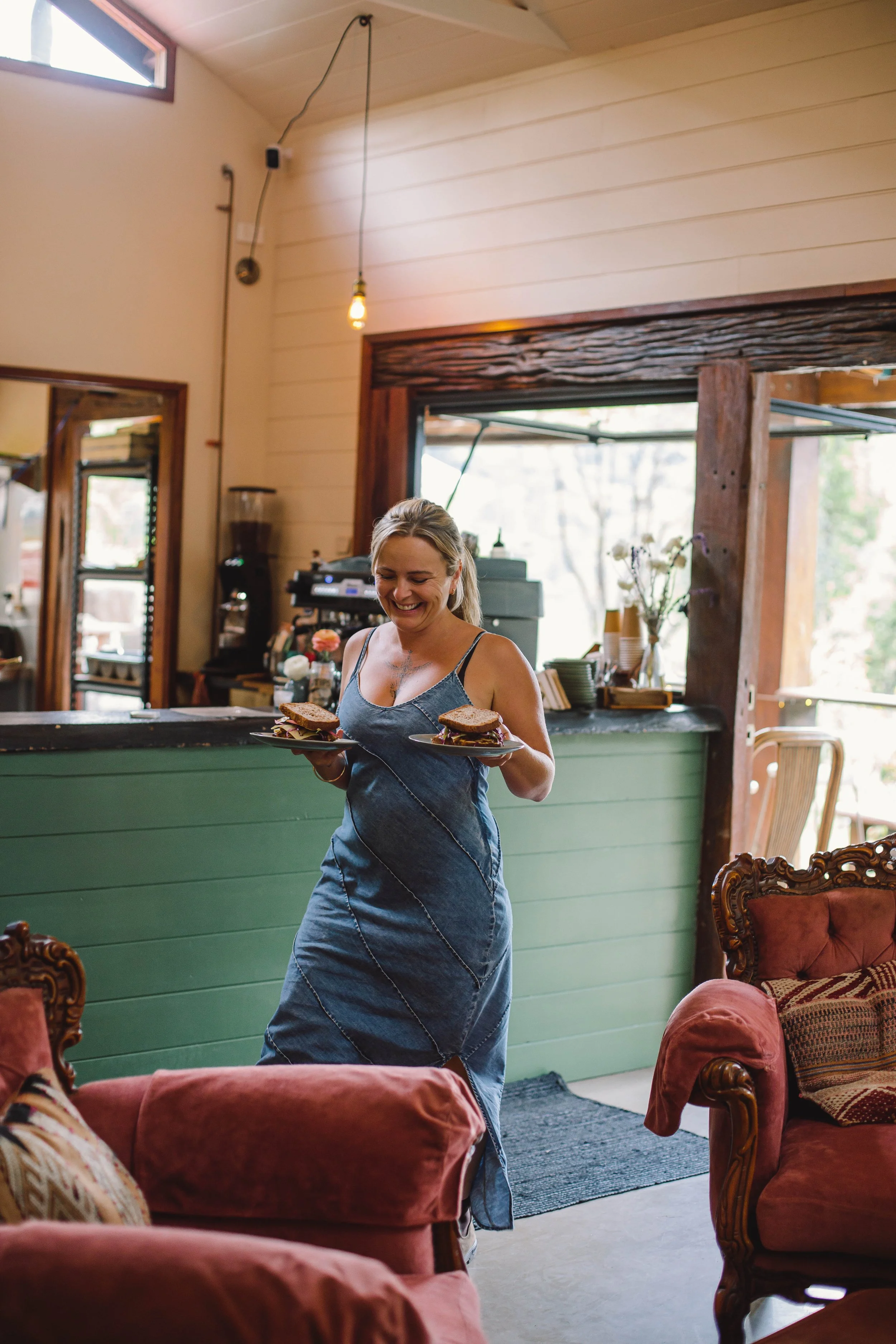 A woman with blond hair smiling and wearing a blue dress carrying two plates with sandwiches in a cozy café or restaurant.