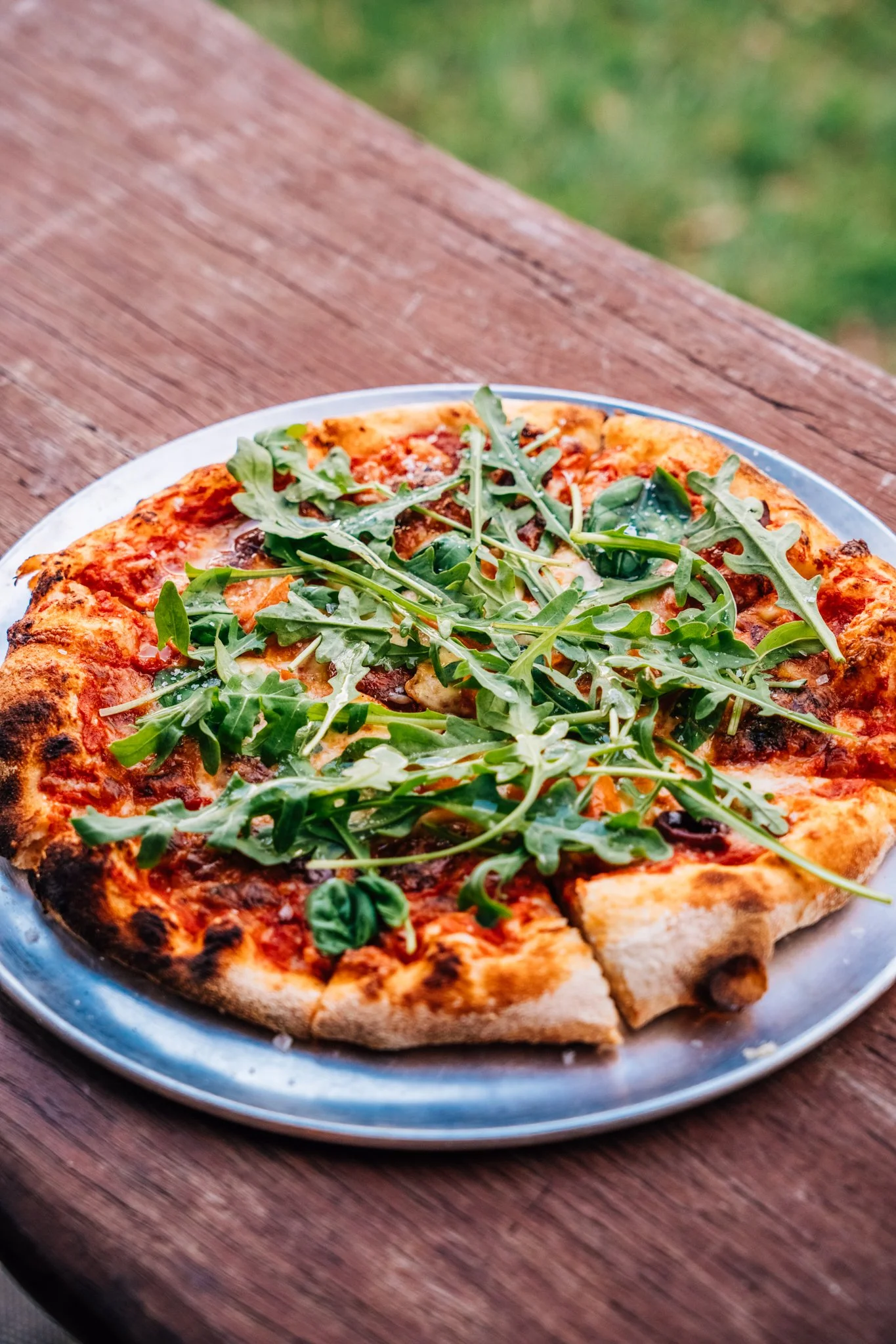 Slice of pizza with arugula on top on a metal plate on a wooden table.