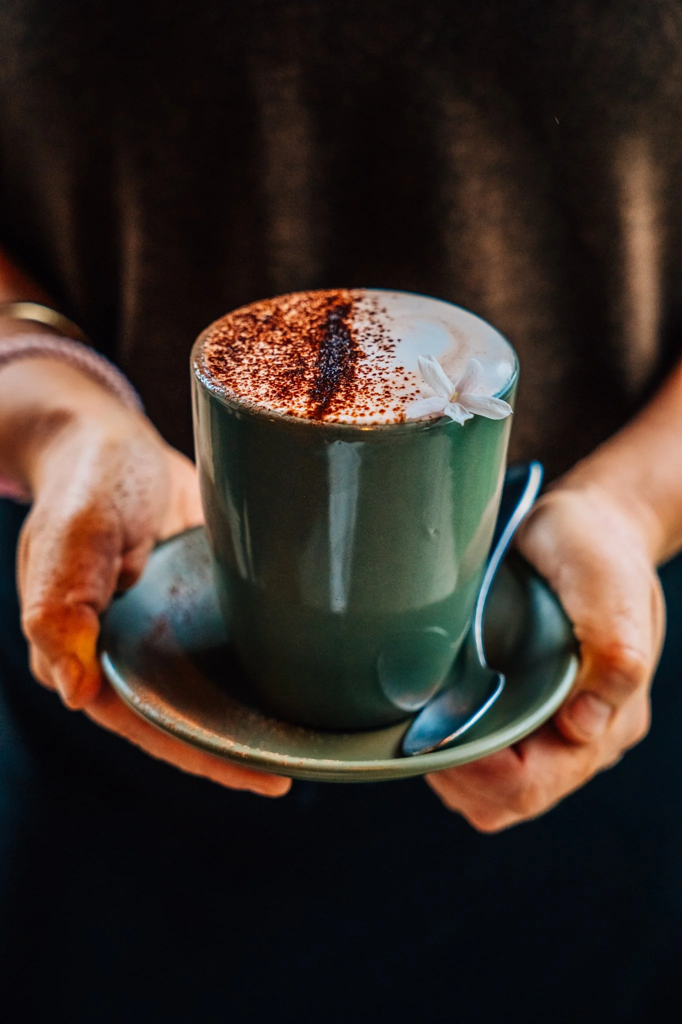 Person holding a green ceramic cup filled with a frothy coffee drink, topped with cocoa powder and a small white flower garnish, on a matching saucer with a spoon.