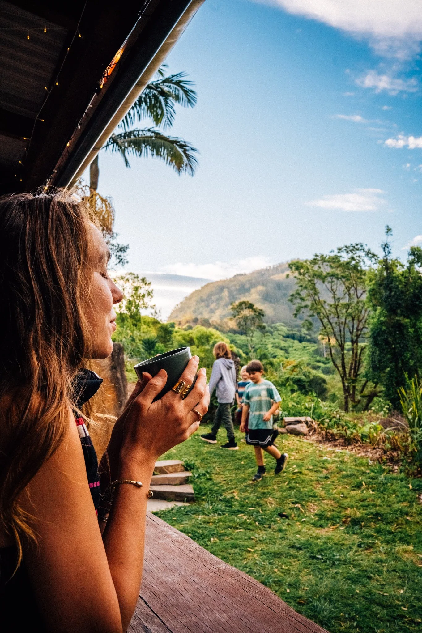 A woman with long auburn hair drinks from a black mug while sitting on a porch, overlooking a green landscape with trees, hills, and a clear blue sky.