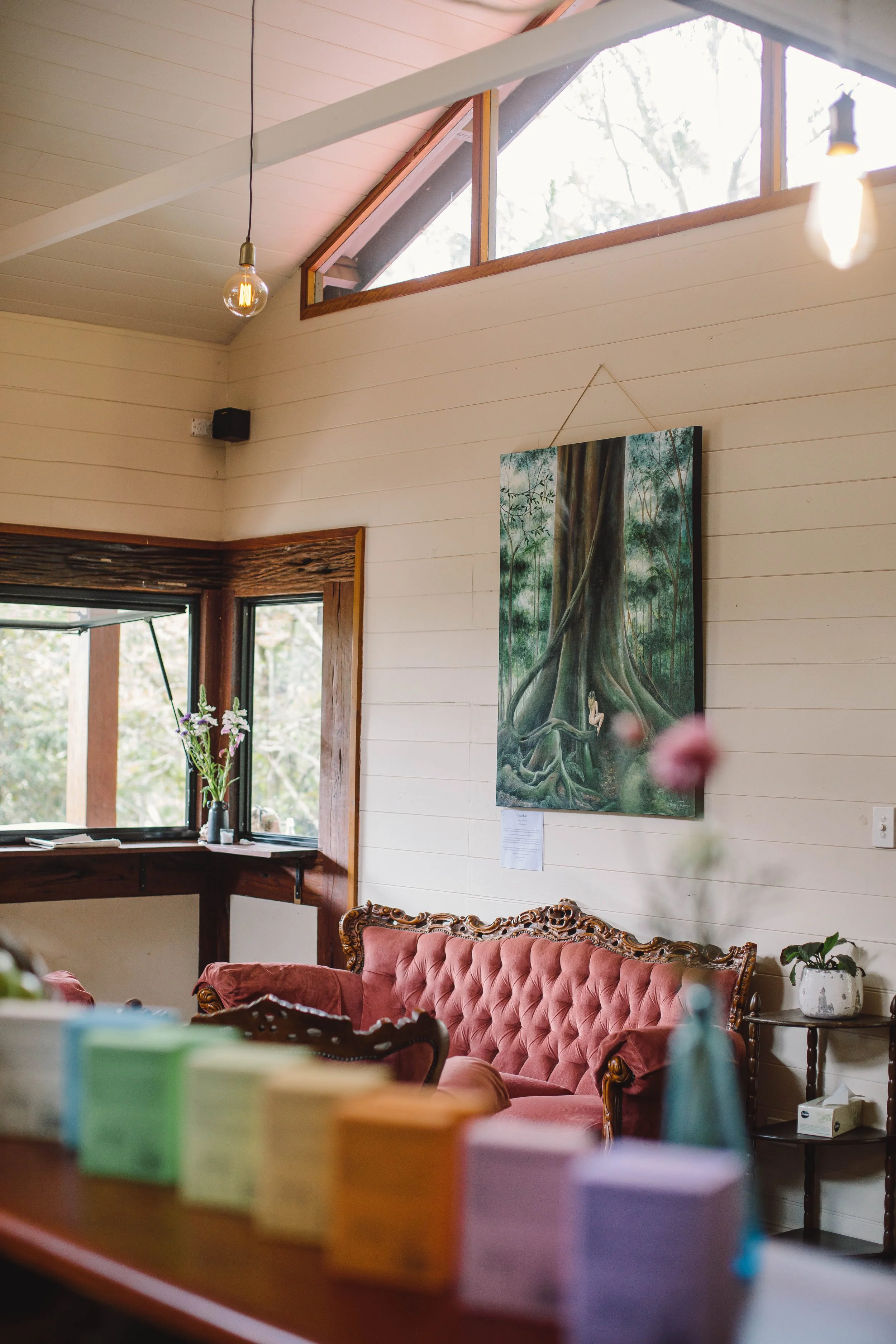 Interior of a cozy room with a vintage pink velvet couch, a wall-mounted painting of a forest scene with a person sitting at the base of a large tree, a window with flowers in a vase on the sill, and soft lighting from hanging bulbs.