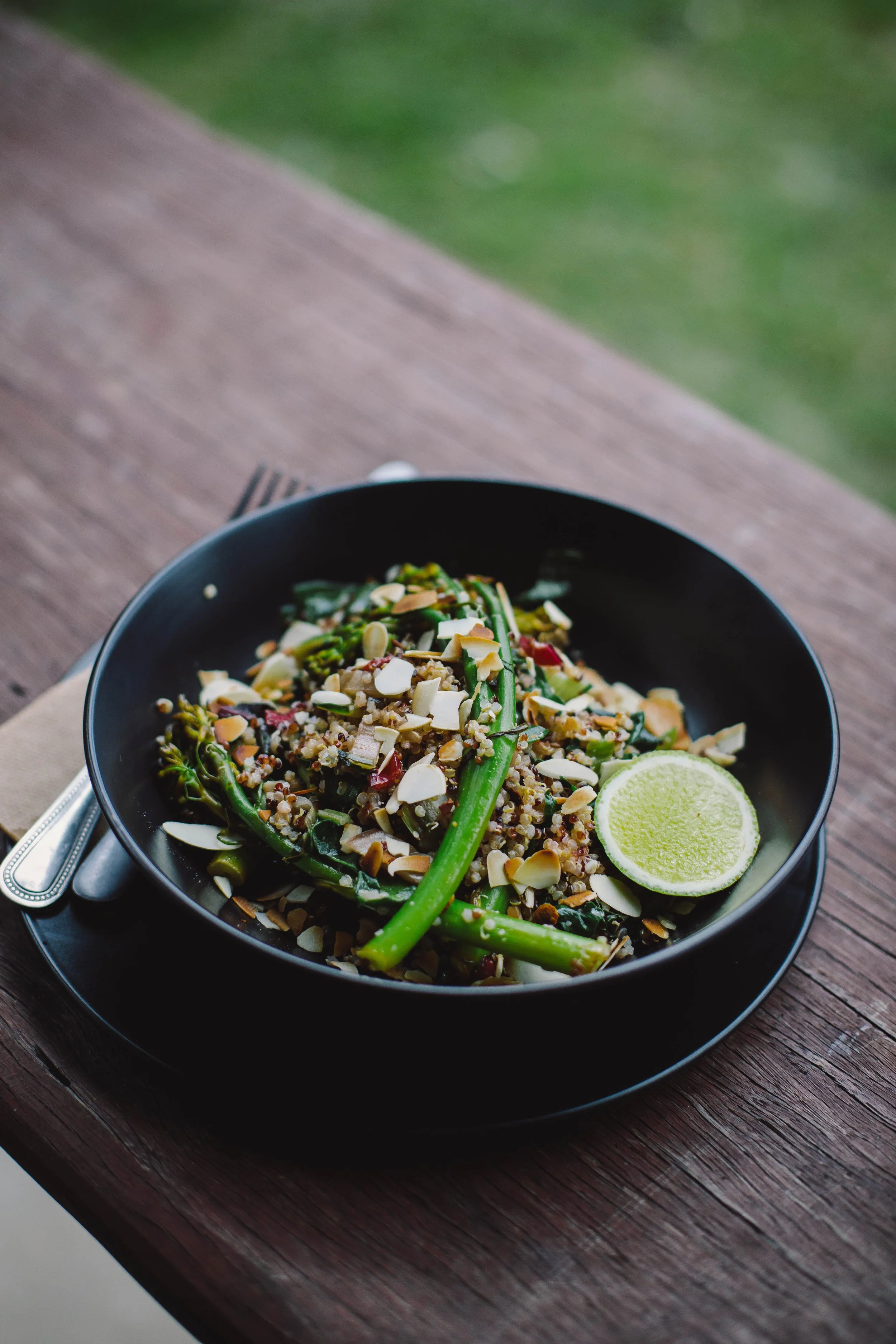 A black bowl of mixed grain salad topped with sliced almonds, green beans, and a lime wedge on a wooden table.