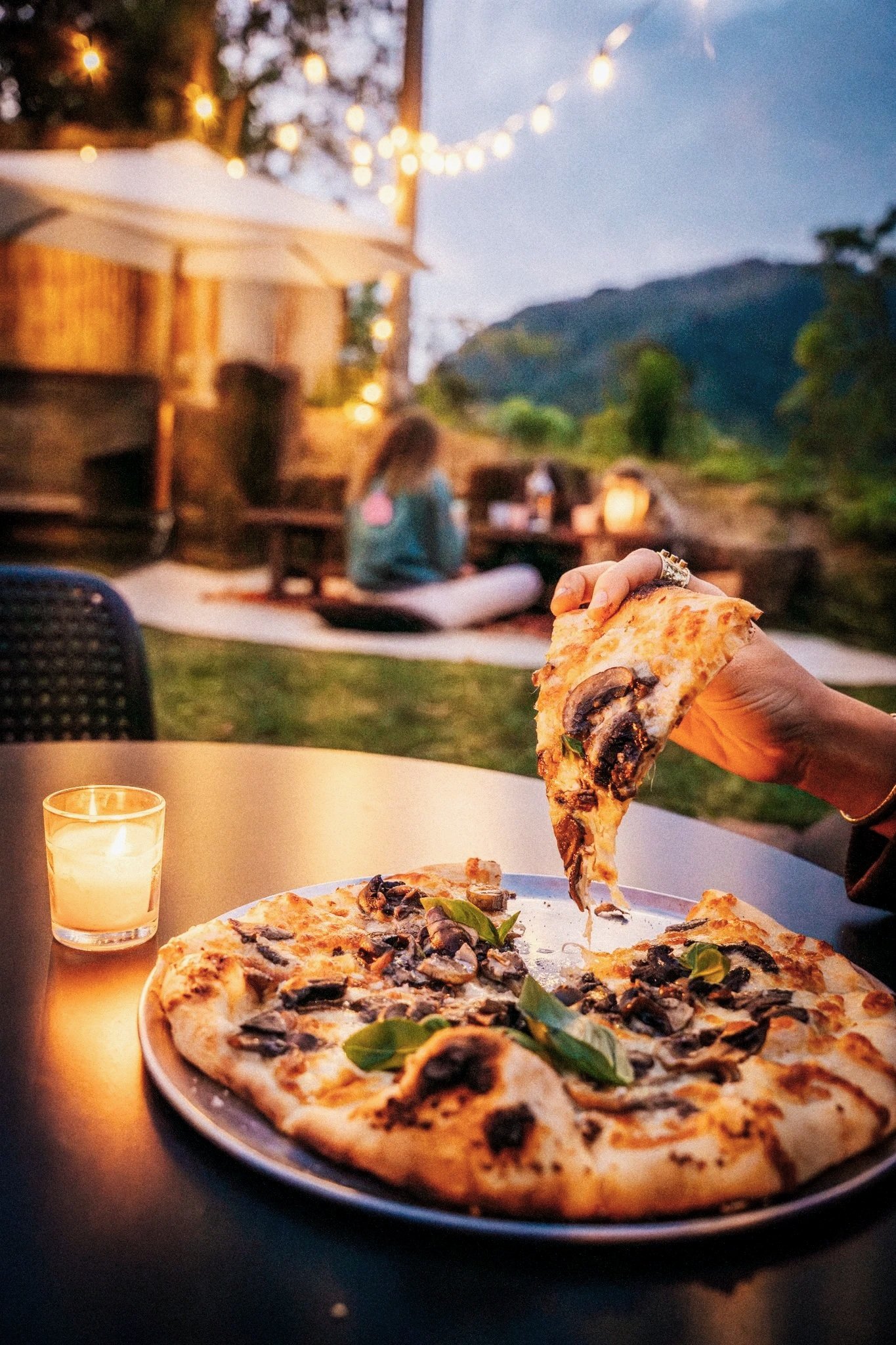 Pizza with mushrooms and basil on a plate, a person lifting a slice, candlelight on a table, outdoor setting with string lights and a mountain backdrop at dusk, people sitting on a blanket in the background.