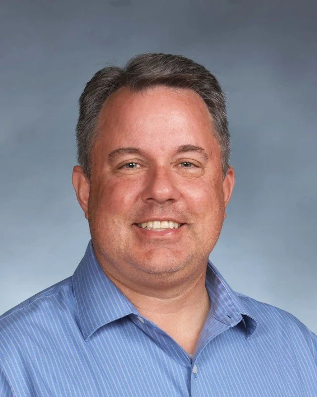 A smiling man with short dark hair, wearing a blue striped dress shirt, against a gray background.