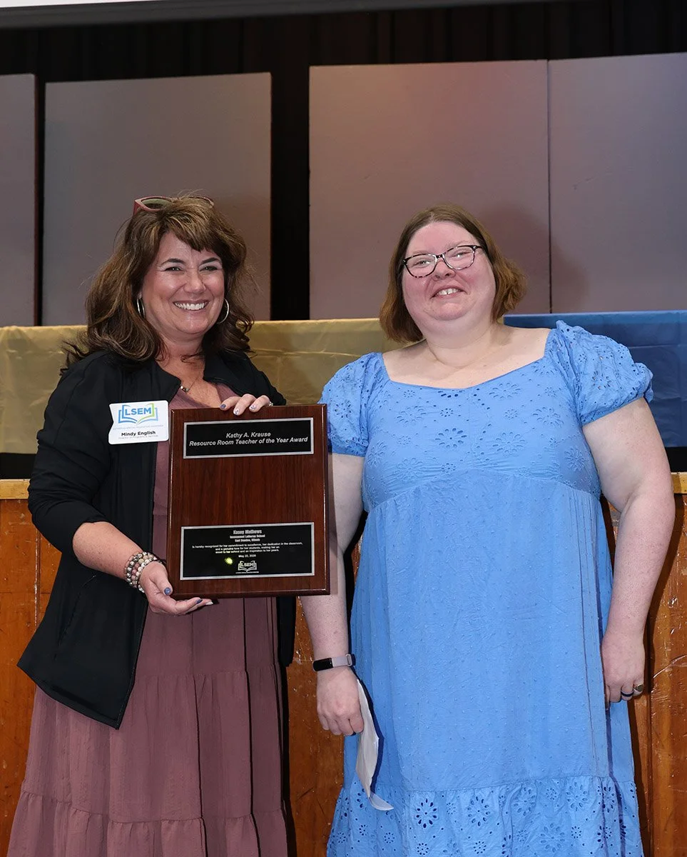 Two women smiling and standing together, one holding a plaque with an award inscription, at an indoor event. The woman on the left is wearing a black jacket and a long pink skirt, with glasses on her head and a name tag. The woman on the right is in a blue dress and glasses, holding a face mask in her hand.