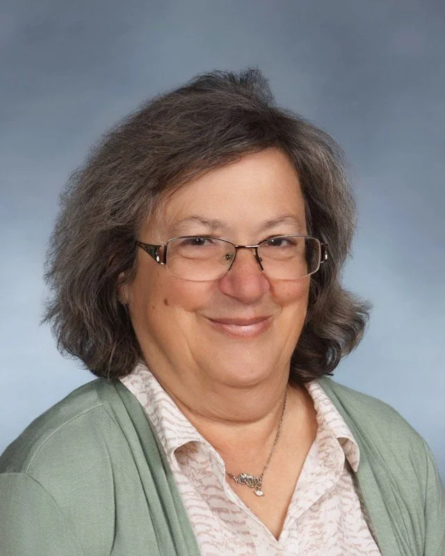 A  woman with glasses and gray hair smiling, wearing a light-colored patterned blouse, a green cardigan, and a necklace, against a blue background.
