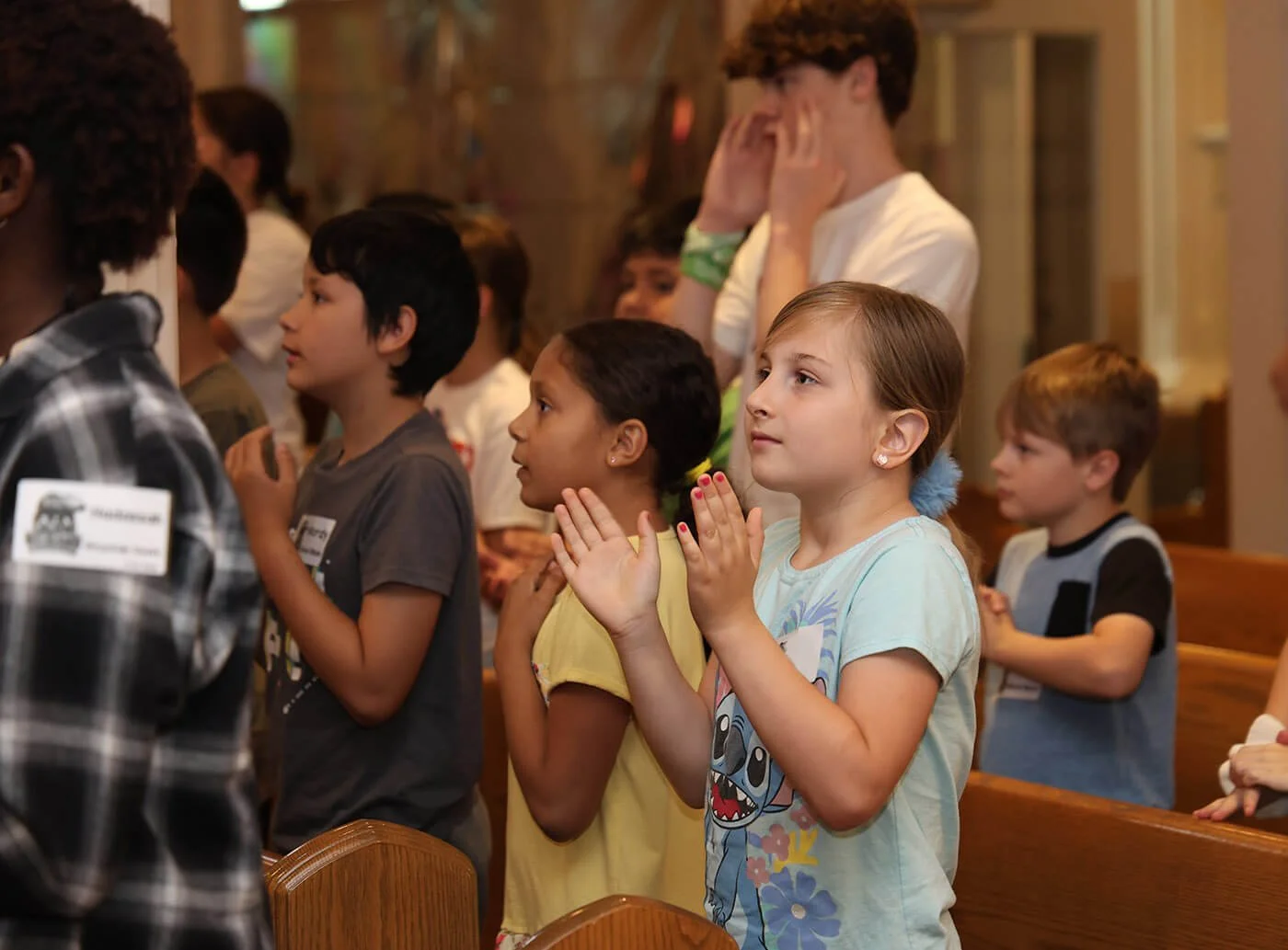 Children and a young adult standing in a church, participating in a prayer or singing activity, with some clapping and some with hands on their hearts.