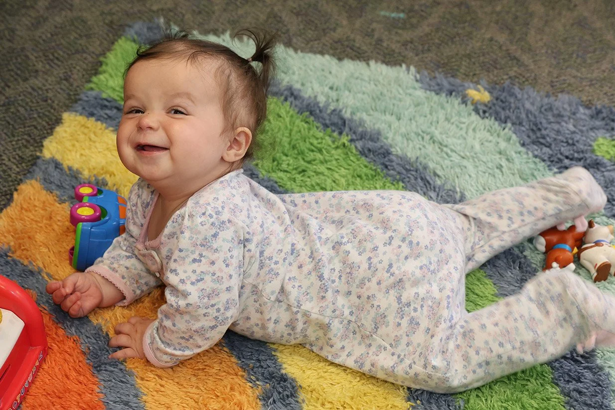 A smiling baby girl lying on a colorful shaggy rug, surrounded by toys, wearing a floral pajamas.