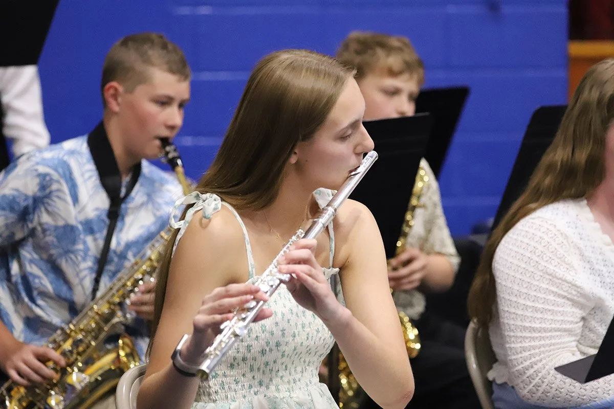 A young woman playing the flute in a school band with other students playing saxophones behind her.