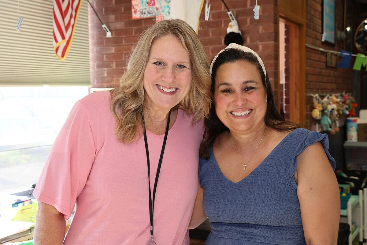 Two smiling women standing close together indoors with a brick wall and colorful decorations in the background.