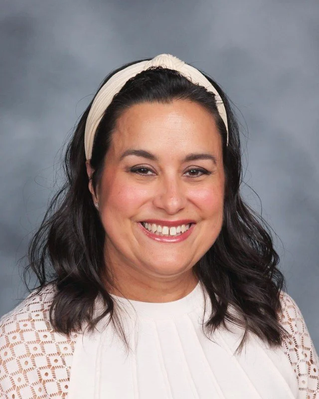 A woman with dark hair, styled in loose waves, wearing a beige headband and a white lace top, smiling at the camera against a gray background.