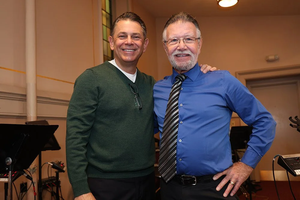 Two men standing indoors, smiling at the camera. One is wearing a green sweater with glasses hanging from the collar, and the other is wearing a blue shirt with a striped tie and glasses, with his hand on his hip.