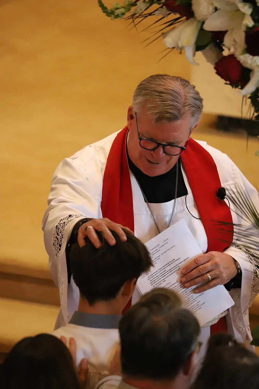 A pastor in a white robe with a red stole and glasses is blessing a boy during confirmation.