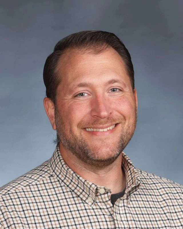 Headshot of a smiling man with brown hair, beard, wearing a plaid shirt, against a neutral gray background.