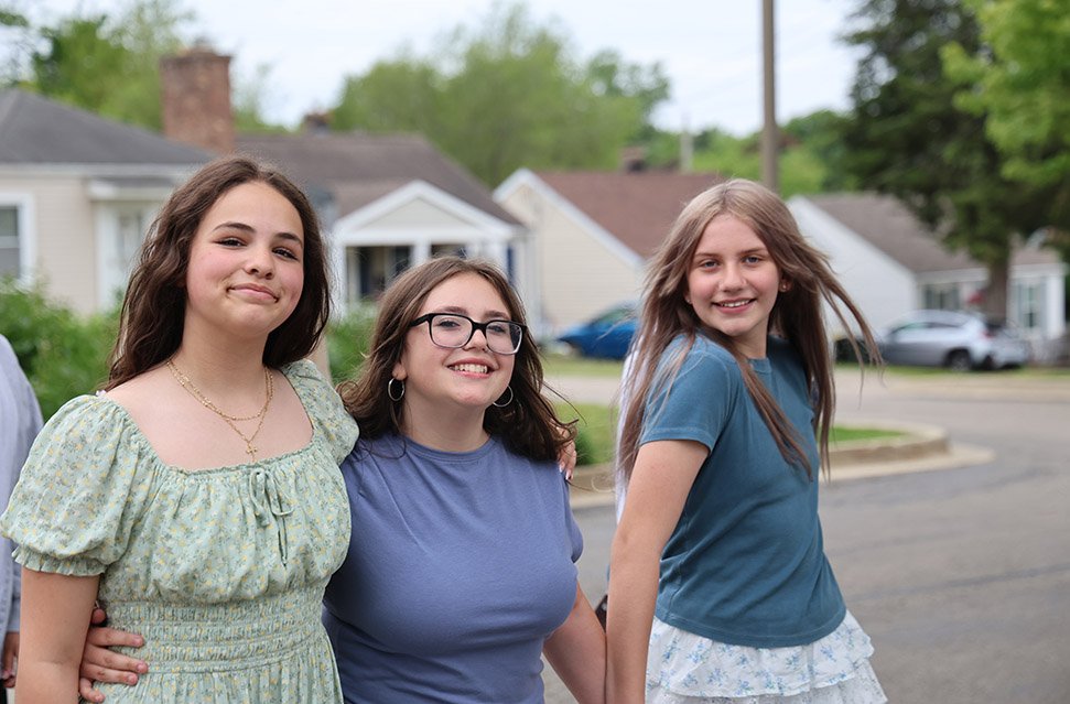 Three young women standing outdoors in a neighborhood with houses and trees in the background, smiling at the camera.