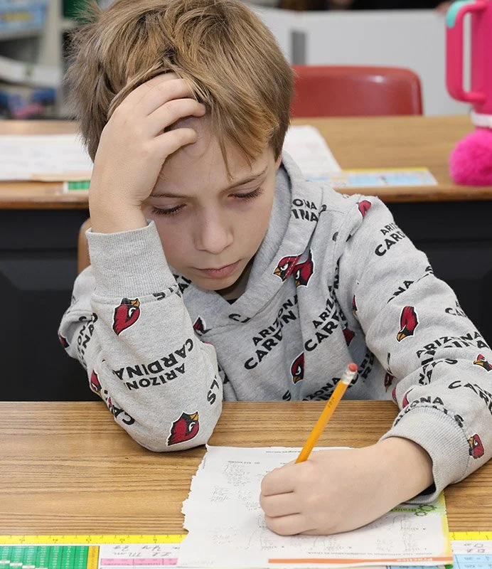 A boy sitting at a desk, holding a pencil, appearing to be focused or frustrated with his homework or writing task.