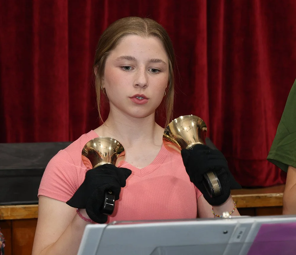 Young woman in pink shirt holding two gold bells, with a red curtain in the background.