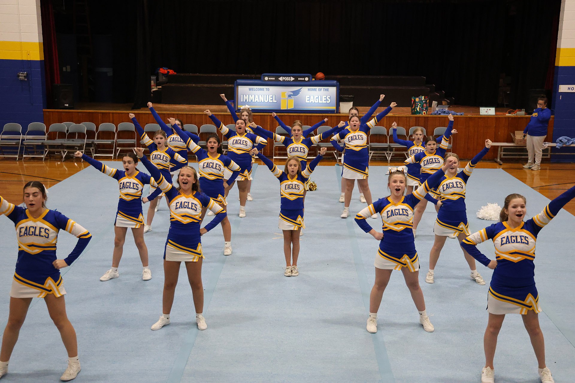 Group of young cheerleaders in blue and yellow uniforms performing a cheer routine on gymnasium floor. Cheering with fists raised, with school sign 'Immanuel Eagles' in background.