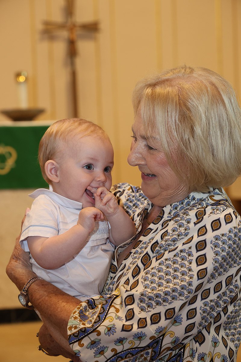 An elderly woman holding a smiling baby boy in her arms, both sharing a joyful moment indoors.
