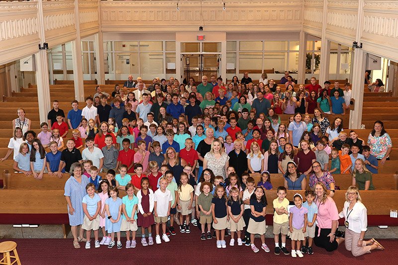 A large group photo of children and adults gathered in a church or auditorium with wooden pews and a high ceiling.