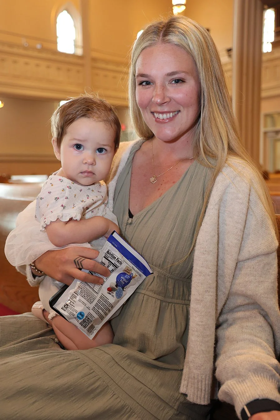 A young woman sitting in a church holding a small girl on her lap. The girl is holding a snack bag. The church has high windows and wooden pews.