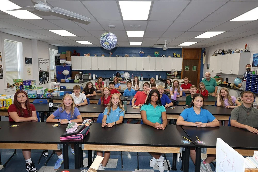 A classroom full of students sitting at desks, with a teacher standing at the back. The room has cabinets, educational decorations, globes, and science supplies.
