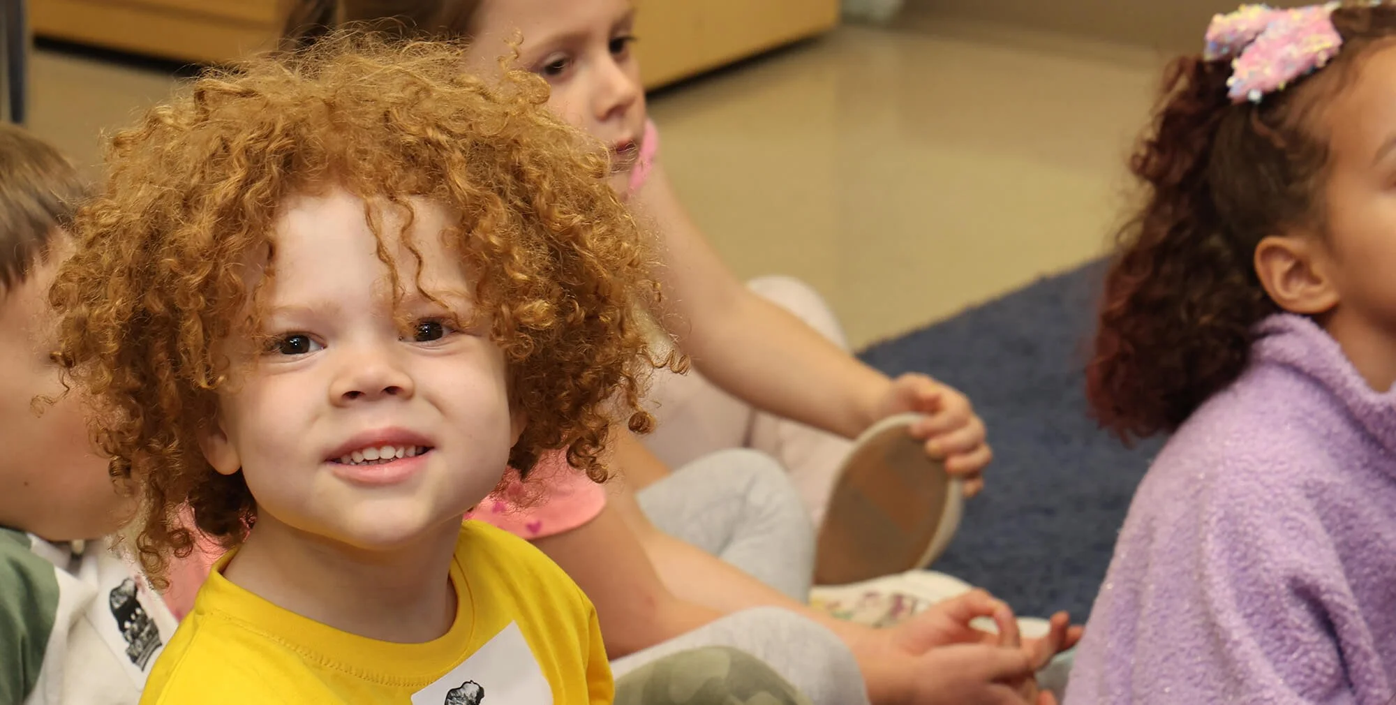 Group of young children sitting on the floor, with one girl with curly red hair and a yellow shirt smiling at the camera, while others are engaged and looking at something off-camera.