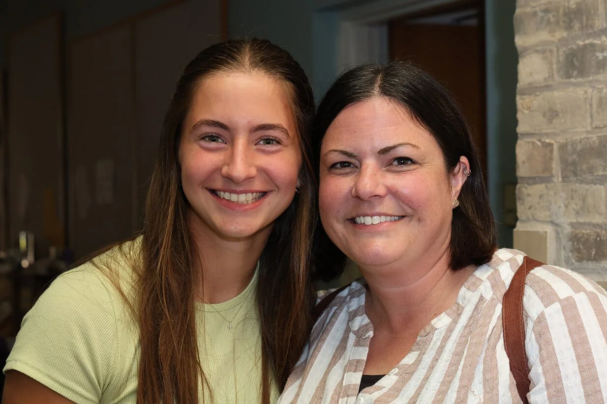 Two smiling women, one younger and one older, posing together indoors near a stone wall.