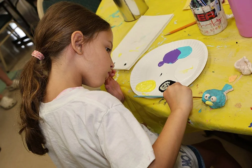 A young girl painting a sculpture of a fish on a yellow table in an art classroom.