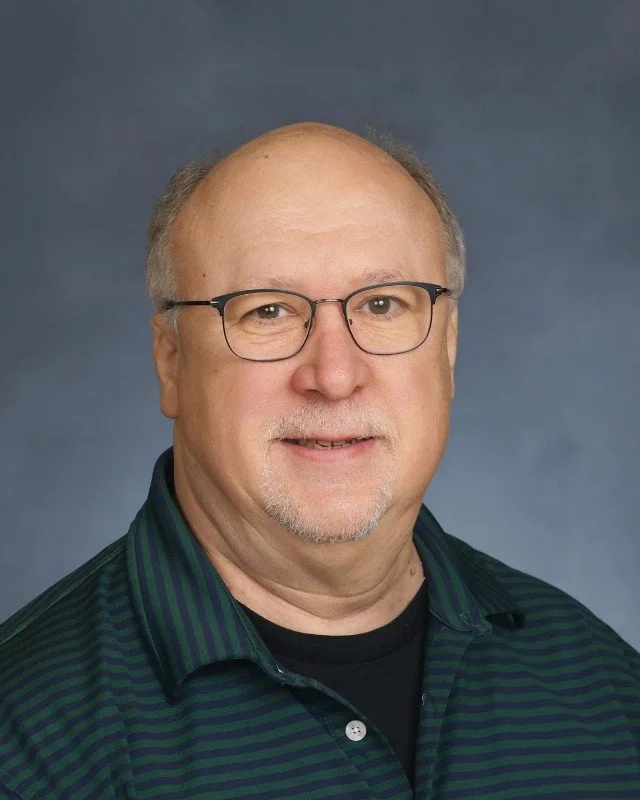 Portrait of a man with glasses, wearing a dark green striped shirt, against a plain blue background.