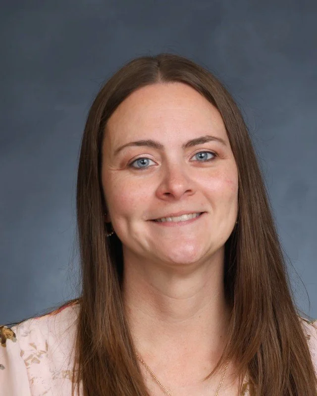 Portrait of a woman with long brown hair, light blue eyes, and a friendly smile, against a blue-gray background.