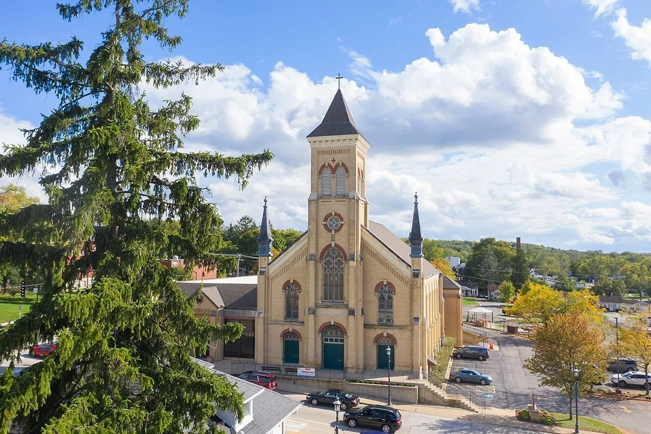A large beige church with a tall steeple and smaller spires sits in a small town, surrounded by parking lots, trees, and some houses, with a partly cloudy sky overhead.