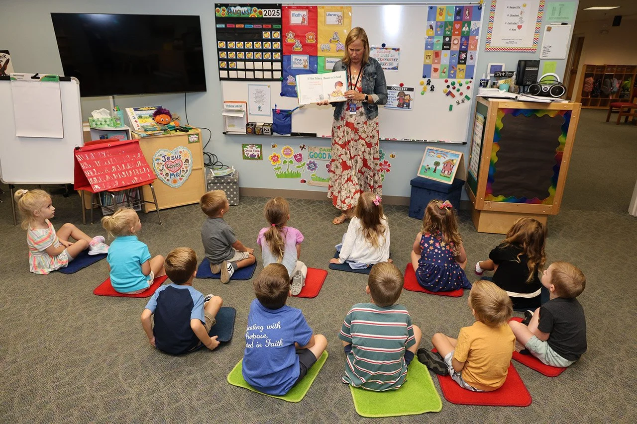 Elementary school classroom with young children sitting on colored mats, listening to a teacher reading a book.