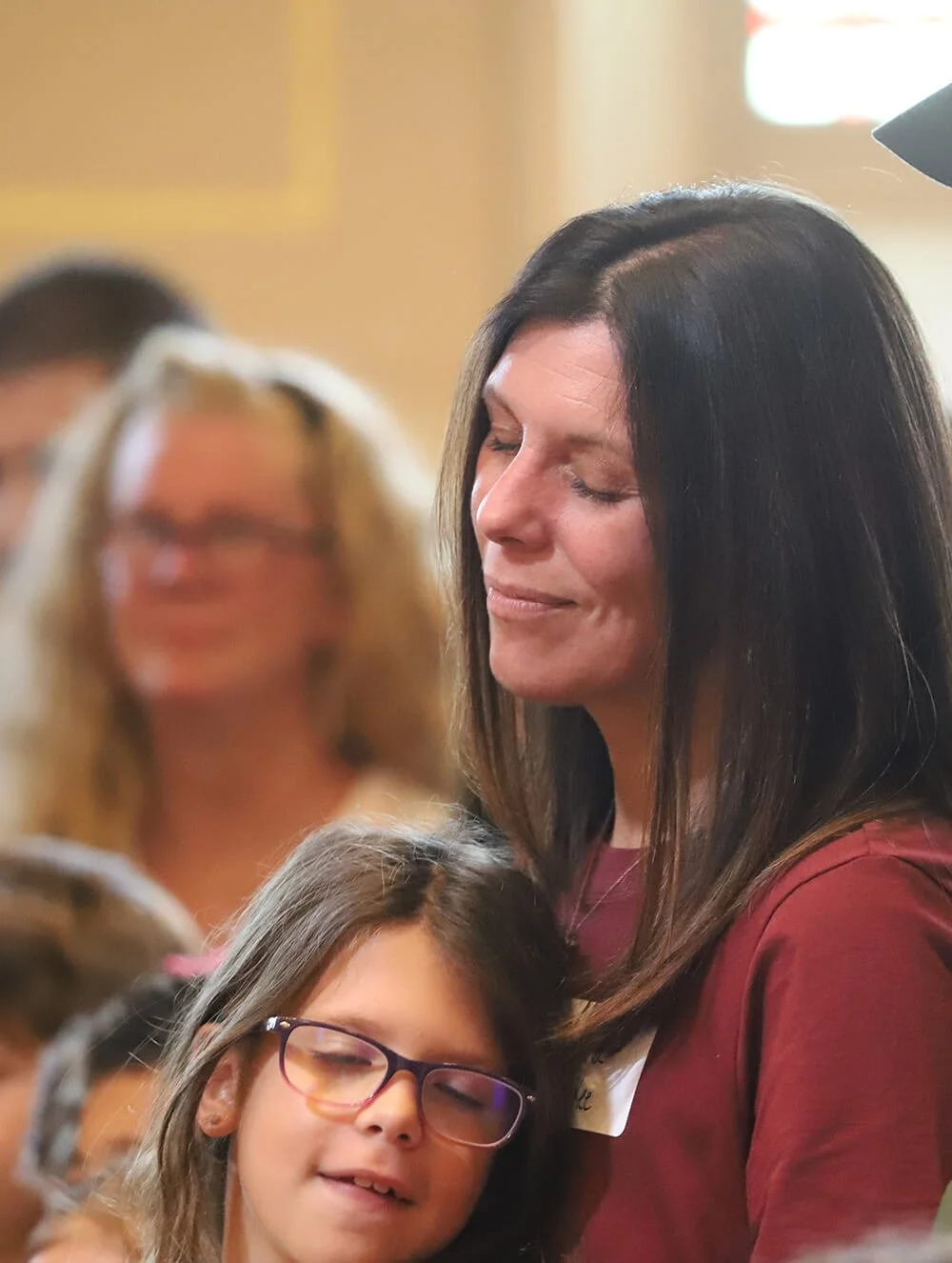 A woman with long dark hair and a maroon shirt is hugging a young girl with glasses and long hair, both with their eyes closed and smiling gently. In the background, other blurred people are visible.