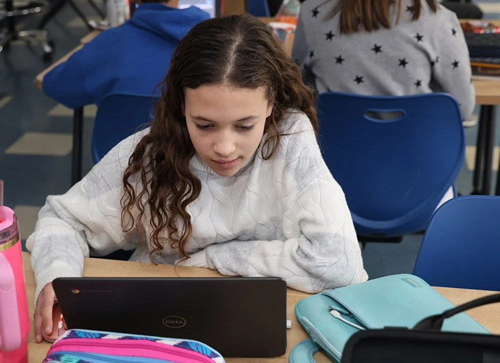 A girl with long curly hair wearing a white sweater, studying on a laptop at a school desk, with a pink water bottle and a light blue pencil case on the desk.